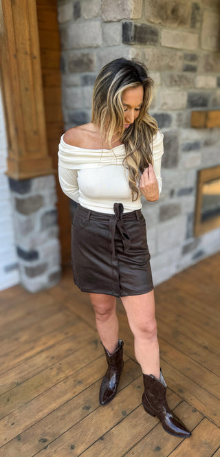 Woman wearing a white off-shoulder top and black leather skirt with brown boots in front of a stone wall.