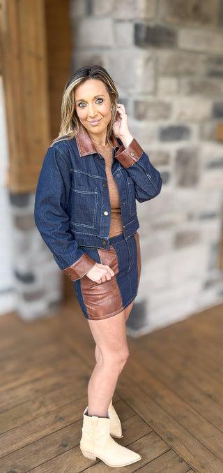 Woman wearing a denim jacket and skirt with brown leather accents indoors.